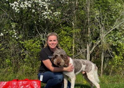 Smiling woman kneeling beside large white and tan spotted poodle in front of bushes